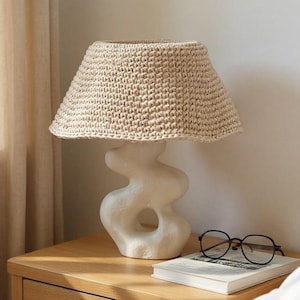 May include: A beige woven lampshade sits atop a white, abstract-shaped lamp base. The lamp is on a wooden nightstand with a book and glasses. The room has neutral tones with a beige curtain in the background. The lamp is a decorative home decor item.