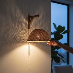 Op de afbeelding: Een bruine, geweven hanglamp is aan een houten beugel bevestigd en werpt een warm licht op de muur. De lampenkap heeft een halfronde vorm. Een hand reikt naar de lamp. Een kleine vaas en boeken liggen op een houten tafel.