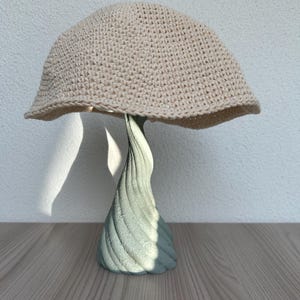 May include: A beige crocheted hat with a wide brim sits atop a light green, spiraled, decorative stand. The hat is made of a tightly woven yarn, and the stand has a textured surface. The hat and stand are on a wooden surface.