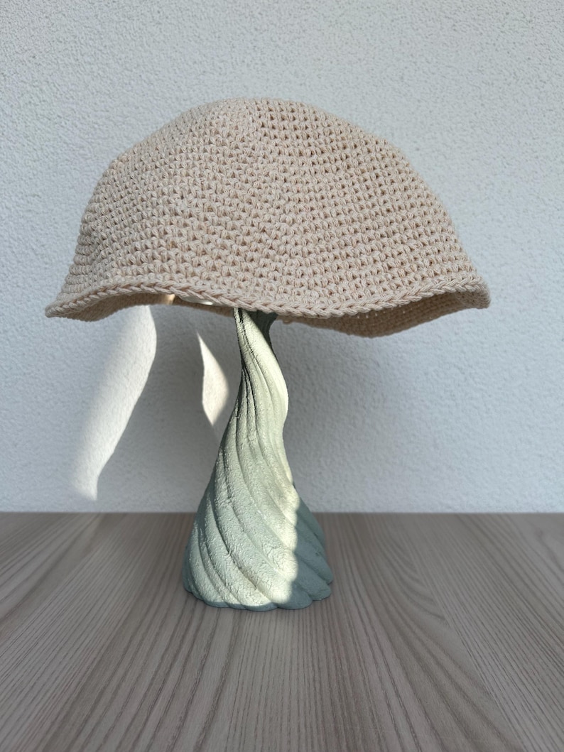 May include: A beige crocheted hat with a wide brim sits atop a light green, spiraled, decorative stand. The hat is made of a tightly woven yarn, and the stand has a textured surface. The hat and stand are on a wooden surface.