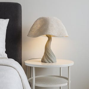 May include: A unique mushroom-shaped table lamp with a textured, off-white crocheted shade. The lamp has a twisted, gray-toned base and sits on a white side table. The lamp is plugged in and illuminated.