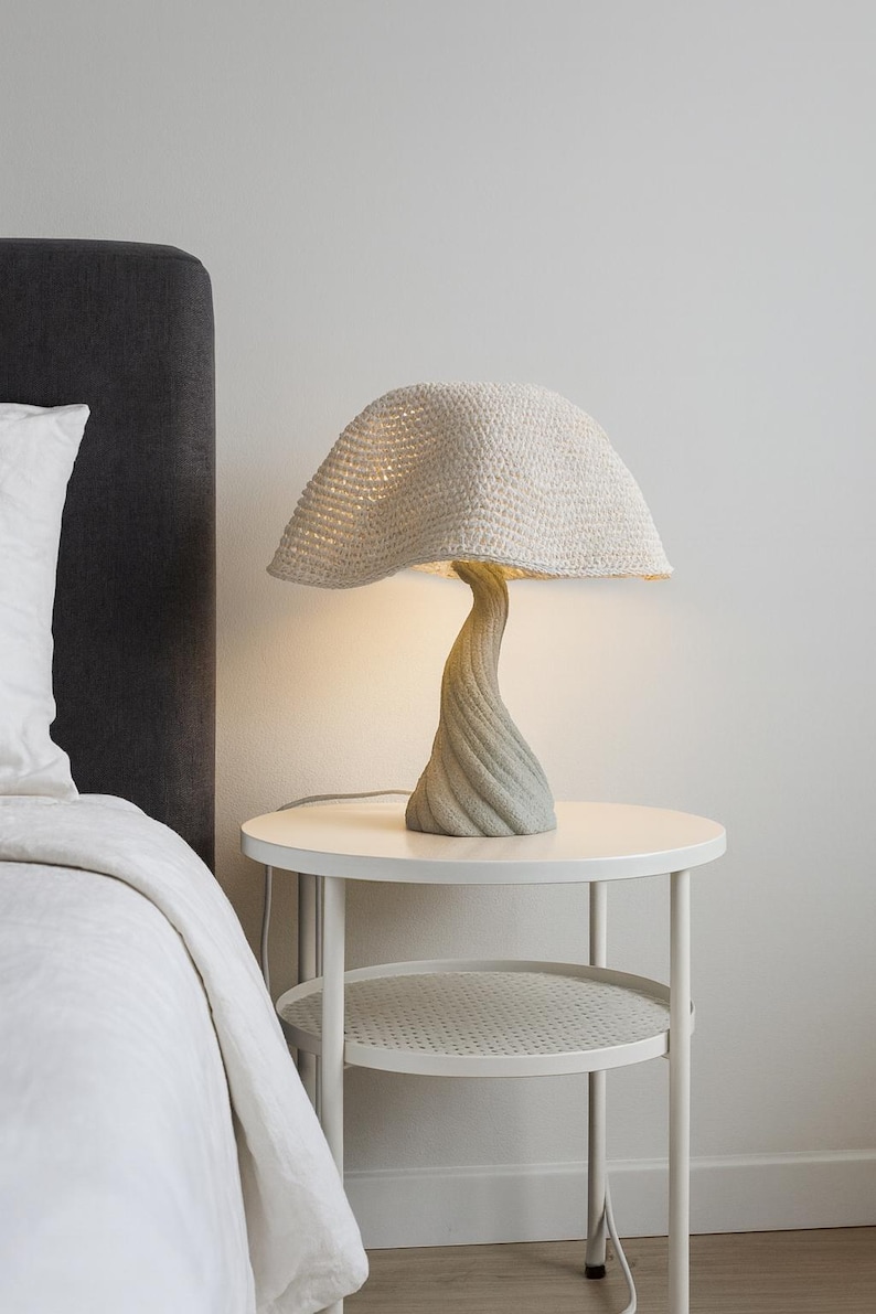 May include: A unique mushroom-shaped table lamp with a textured, off-white crocheted shade. The lamp has a twisted, gray-toned base and sits on a white side table. The lamp is plugged in and illuminated.