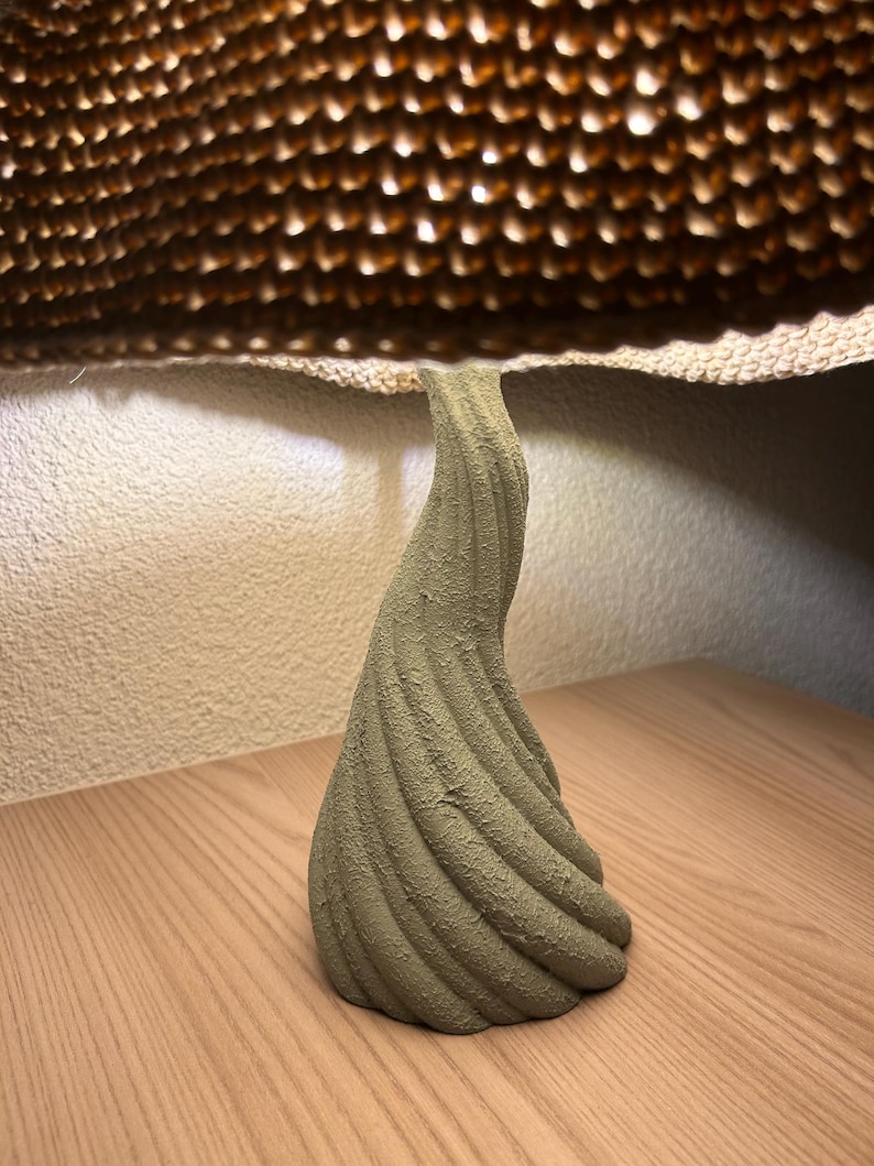 May include: A textured, olive-green, abstract sculpture with a spiral design. The sculpture is on a wooden surface, with a textured, brown, honeycomb-like structure above it.