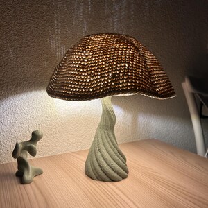 May include: A mushroom-shaped lamp with a textured, woven lampshade and a twisted, light green base. The lamp is illuminated, casting a warm glow. A small, abstract sculpture sits nearby. The lamp is on a wooden surface.