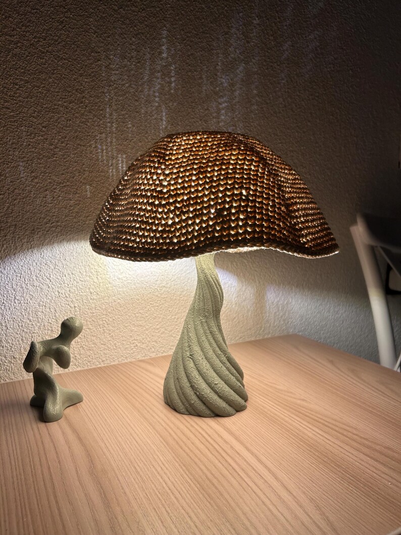 May include: A mushroom-shaped lamp with a textured, woven lampshade and a twisted, light green base. The lamp is illuminated, casting a warm glow. A small, abstract sculpture sits nearby. The lamp is on a wooden surface.