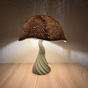 Handmade mushroom lamp with a twisted sculptural base and handwoven brown shade. Emits a warm, cozy light that creates soft shadows on the wall, perfect for nightstands or ambient decor.