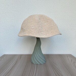 May include: A beige crocheted hat with a rounded crown and a slightly downward-sloping brim. The hat is displayed on a light green, twisted, decorative stand. The stand is on a wooden surface.
