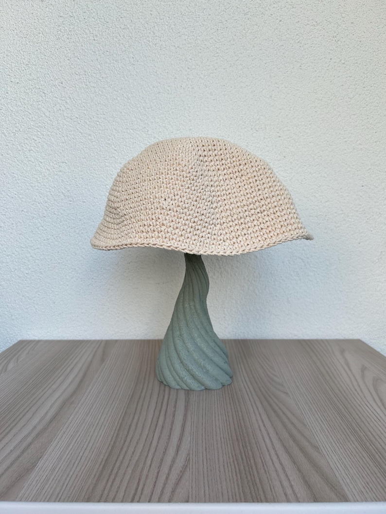 May include: A beige crocheted hat with a rounded crown and a slightly downward-sloping brim. The hat is displayed on a light green, twisted, decorative stand. The stand is on a wooden surface.