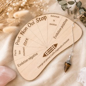 Puede incluir: Una tabla de radiestesia de madera en forma de semicírculo con texto grabado en francés que incluye "Oui", "Non", "Stop" y "Evolution positive", junto con un péndulo de piedra marrón en punta sobre una cadena de metal plateado.
