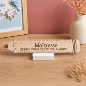 Peut inclure: Une règle en bois clair en forme de crayon, avec une pointe et une gomme gravées en brun foncé. Le texte "Maîtresse" et "MERCI POUR CETTE BELLE ANNÉE" est gravé au centre. Elle repose sur un petit socle rectangulaire de couleur blanche.