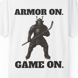 May include: White t-shirt with the text "ARMOR ON. GAME ON." above and below a graphic of a samurai in armor holding a hockey stick. The samurai is in a battle stance.