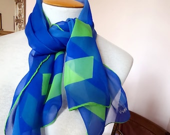 May include: A blue and green patterned silk scarf with the word "Usma" printed on the end. The scarf is tied around a white mannequin's neck.