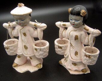 May include: Two ceramic figurines of people carrying baskets on a yoke. The figurines are painted in pink and gold with a white snow-like texture. The figures are wearing traditional Asian clothing.