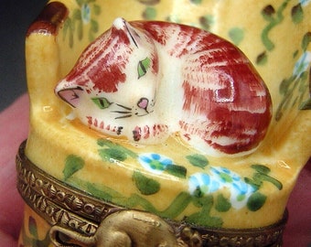 May include: A porcelain trinket box shaped like a yellow armchair with a red and white striped cat sleeping on it. The chair is decorated with green leaves and blue flowers. A gold mouse is on the side of the box.