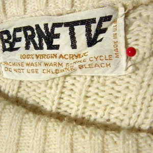 May include: White knit sweater with a white tag that reads "BERNETTE 100% VIRGIN ACRYLIC MACHINE WASH WARM GENTLE CYCLE DO NOT USE CHLORINE BLEACH MADE IN U.S."