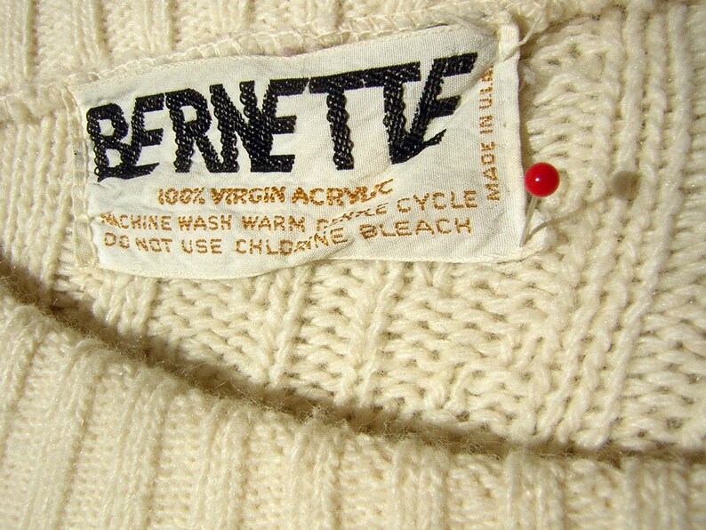 May include: White knit sweater with a white tag that reads "BERNETTE 100% VIRGIN ACRYLIC MACHINE WASH WARM GENTLE CYCLE DO NOT USE CHLORINE BLEACH MADE IN U.S."