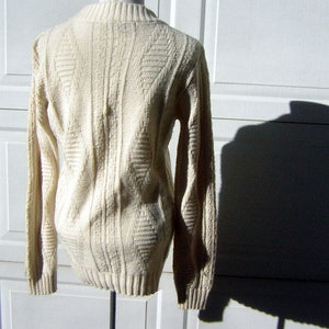 May include: A cream-colored knitted sweater with a cable knit pattern. The sweater has long sleeves and a crew neck.