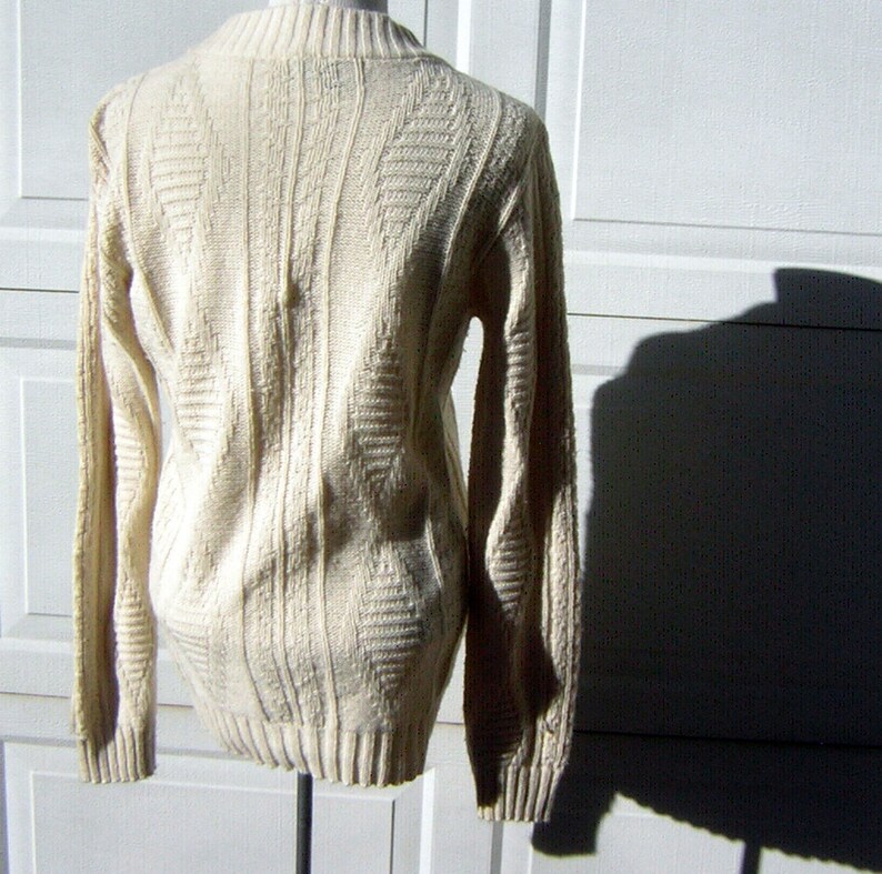 May include: A cream-colored knitted sweater with a cable knit pattern. The sweater has long sleeves and a crew neck.
