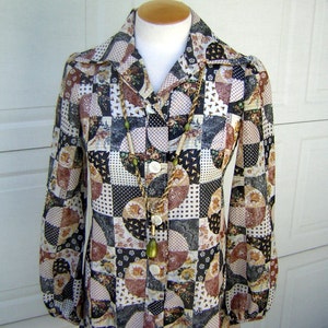 May include: A long-sleeved blouse with a patchwork design featuring various shades of brown, black, white, and cream. The fabric has a geometric pattern with circles, squares, and triangles. The blouse has a button-up front and a collar. A long, beaded necklace with a pendant hangs from the collar.