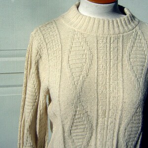 May include: A cream-colored knitted sweater with a cable knit pattern. The sweater has a high neck and long sleeves.