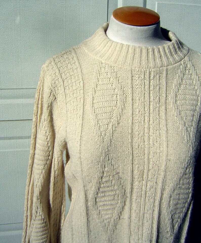 May include: A cream-colored knitted sweater with a cable knit pattern. The sweater has a high neck and long sleeves.