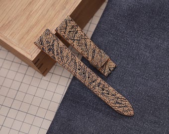 Könnte beinhalten: Zwei Uhrenarmbänder mit beigem und schwarzem strukturiertem Muster, auf blauem Stoff und einer Holzbox. Die Armbänder haben ein reptilienartiges Aussehen.
