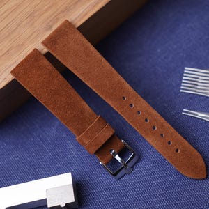 Può includere: Due cinturini per orologio in pelle scamosciata marrone con fibbia argentata e fori di regolazione multipli. I cinturini sono esposti su una superficie blu, con una scatola di legno e strumenti metallici sullo sfondo. I cinturini sono marrone caldo.
