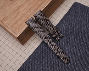 Könnte beinhalten: Zweiteiliges Uhrenarmband mit Schlangenmuster. Das Armband ist überwiegend grau mit schwarzen Akzenten und hat eine strukturierte Oberfläche. Das Uhrenarmband liegt auf einem blauen Stoff neben einer Holzkiste.