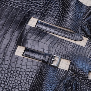 May include: Close-up of black alligator leather watch straps and matching leather. The leather has a textured, scale-like pattern. The watch straps have silver buckles and are laid out on a grid.