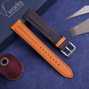 May include: Two watch straps, one orange and one black, with silver buckles, are displayed on a blue surface. The orange strap has black stitching, and the black strap has orange stitching. Tools and a spool of thread are also visible.