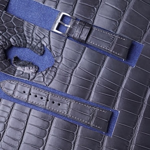Può includere: Primo piano di cinturini per orologi in pelle di coccodrillo grigio scuro. I cinturini sono posizionati su uno sfondo di tessuto blu. Un cinturino ha una fibbia argentata. La pelle ha un motivo strutturato a scaglie.