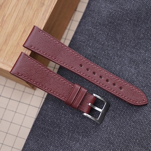 Peut inclure: Une paire de bracelets de montre bordeaux avec une surface texturée et des bords cousus. Les bracelets ont une boucle argentée et sont présentés sur un tissu gris et une boîte en bois.