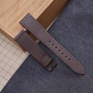 May include: A brown leather watch strap with contrasting stitching. The strap is made of two pieces, one with holes for the buckle and the other with a loop. The watch strap is shown on a textured surface.