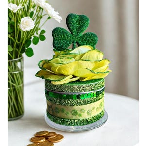 May include: A decorative St. Patrick's Day centerpiece featuring a green and yellow floral arrangement. The centerpiece includes a shamrock, glitter, and a ribbon design. Gold coins are scattered on the table, and a vase of white flowers is in the background.