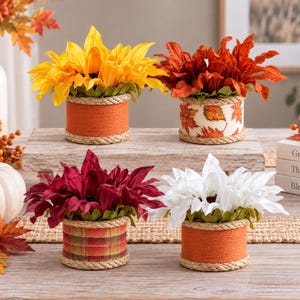 May include: Four decorative flower arrangements in cylindrical containers wrapped in rope. The pots feature orange fabric, a white leaf print, a red plaid pattern, and orange fabric. Each holds bright yellow, rust, maroon, or white flowers.