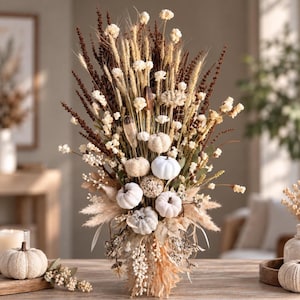 May include: A tall floral arrangement featuring dried wheat stalks, white flowers, and small decorative pumpkins in shades of white and cream. The bouquet includes dark brown feathery plumes and textured botanical elements, creating a dense, vertical display of natural, dried plant materials.