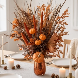 May include: A decorative floral arrangement in a copper-colored glass vase featuring orange pumpkins, brown pinecones, dried grasses, and autumn-toned foliage. The centerpiece is displayed on a table with white candles and small acorns nearby.