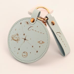 Könnte beinhalten: Ein hellblaues, rundes Schlüsselanhänger mit einem goldenen Ring und Riemen. Der Schlüsselanhänger zeigt ein Krebs-Sternzeichen-Design mit einer Krabbe, Planeten und Sternen. Das Wort "Krebs" steht auf dem Schlüsselanhänger und dem Riemen.