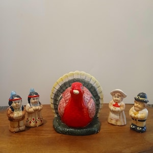 May include: Thanksgiving-themed ceramic figurines. A large red turkey is surrounded by smaller figures of Native Americans, a pilgrim woman, and a pilgrim man. The figurines are painted with bright colors and detailed features, perfect for holiday decor.