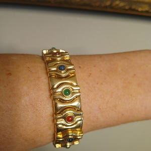 Puede incluir: Un brazalete dorado con eslabones rectangulares, cada uno con pequeñas piedras preciosas redondas en rojo, verde y azul. El brazalete se lleva en una muñeca.