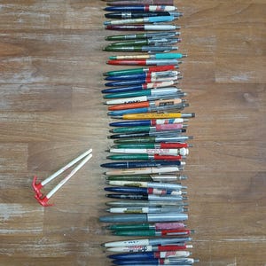 May include: A collection of ballpoint pens in various colors, arranged vertically on a wooden surface. The pens have different colored barrels, including red, blue, green, and silver. Some pens have text printed on them. Two white pens with red and white accents are also present.