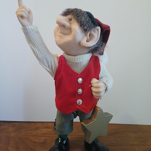 Op de afbeelding: Een grillig elfje met een rood vest, groene broek en zwarte schoenen. De elf houdt een gouden sterornament vast en wijst omhoog. De elf heeft een witte trui en een rood-groene muts.