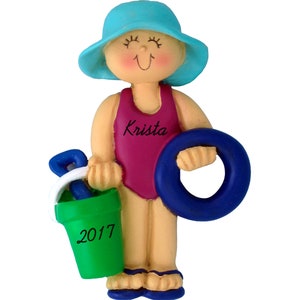 May include: A personalized Christmas ornament featuring a girl wearing a blue hat and a pink swimsuit. She is holding a blue inflatable ring and a green bucket with a blue shovel. The ornament is inscribed with the name "Krista" and the year "2017".