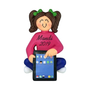 May include: A personalized Christmas ornament featuring a girl with brown hair and pigtails wearing a pink sweater and blue jeans. She is holding a tablet with a blue screen and the text "Mandi 2019" is printed on her sweater.