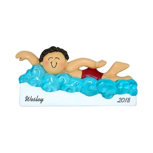 May include: A personalized Christmas ornament featuring a cartoon swimmer in a red swimsuit, swimming in blue water. The ornament has a white base with the name "Wesley" and the year "2018" printed on it.