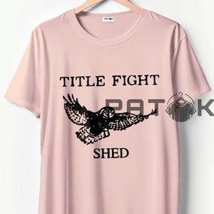 May include: A pale pink t-shirt featuring the text "TITLE FIGHT SHED" and a black owl graphic. The t-shirt is displayed on a wooden hanger.