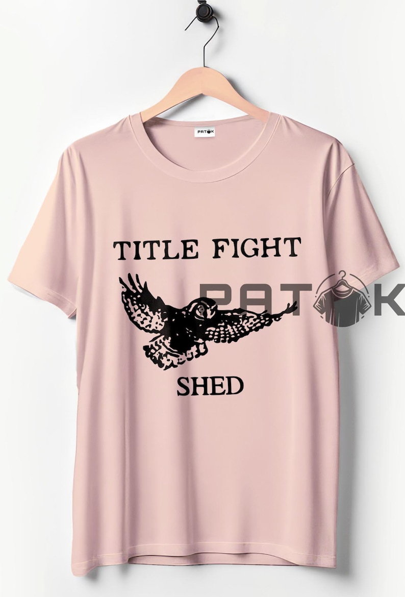 May include: A pale pink t-shirt featuring the text "TITLE FIGHT SHED" and a black owl graphic. The t-shirt is displayed on a wooden hanger.