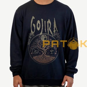 May include: Navy blue sweatshirt featuring the band name "GOJIRA" in a distressed gold font. The front graphic includes a circular design with a tree, moon, and other symbols. The sweatshirt is long-sleeved.