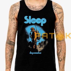 Pu&ograve; includere: Canotta nera con il nome della band "Sleep" in blu, sopra un'illustrazione di una figura con uno zaino in un paesaggio arancione e blu. La parola "dopesmoker" &egrave; stampata sotto.