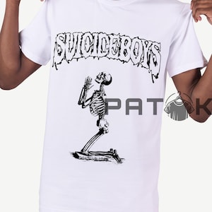 May include: White t-shirt with the text "SUICIDEBOYS" above a black and white illustration of a kneeling skeleton in prayer. The design is centered on the front of the shirt.